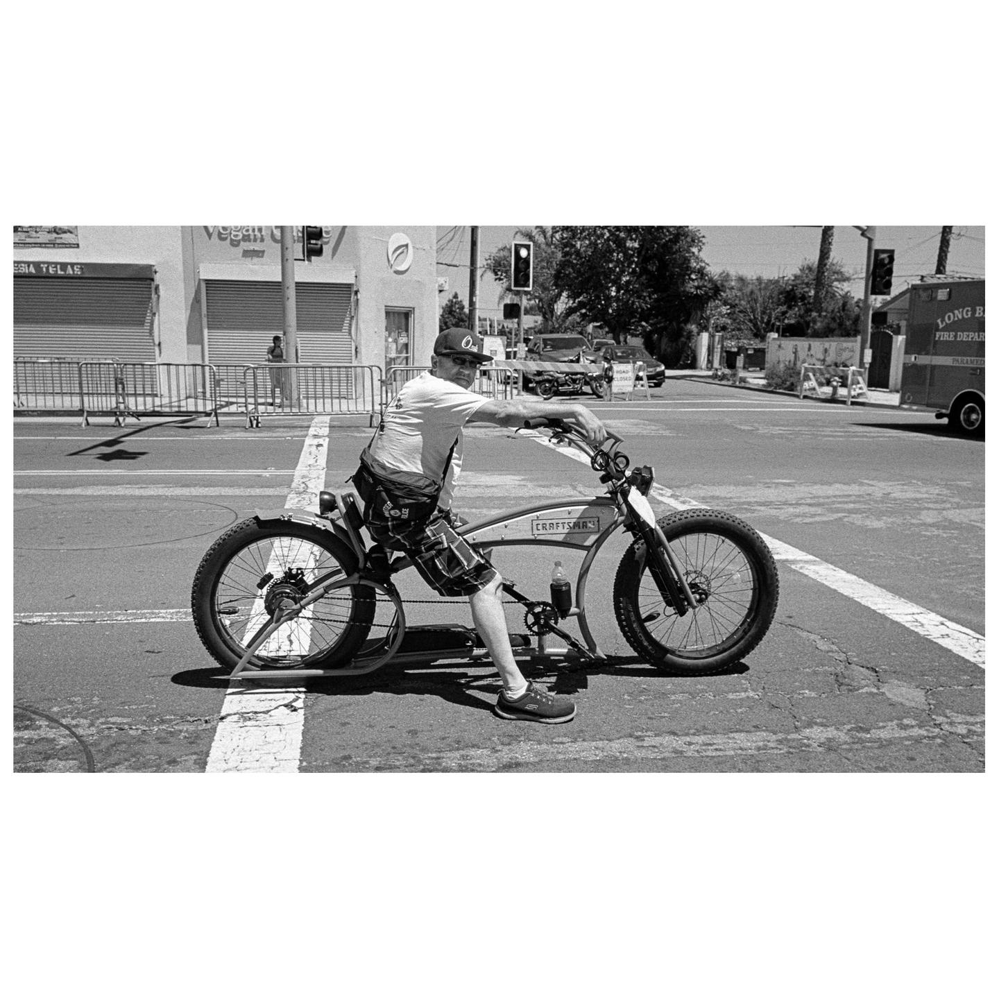 A man sits on a bicycle in the middle of a street with buildings and an emergency vehicle in the background.
