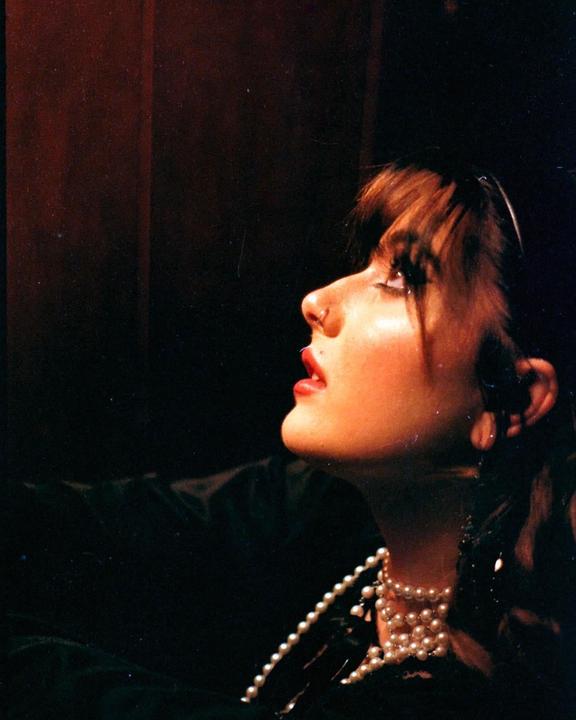 A woman with bangs and a pearl necklace looks up, illuminated by a light source from above.