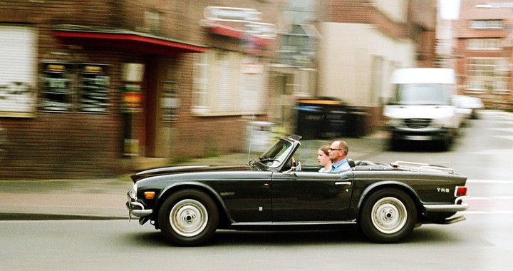 A man and a girl ride in a convertible car on a street with blurred buildings in the background.