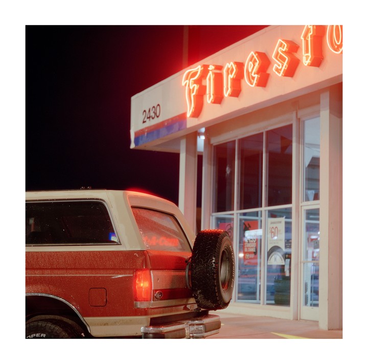 A red and white vehicle with a spare tire on the back is parked at night in front of a building with a neon sign.