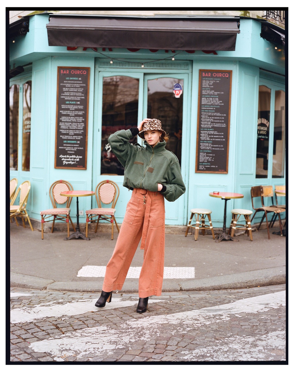 A woman wearing a leopard print hat and a green jacket stands on a street in front of a cafe.