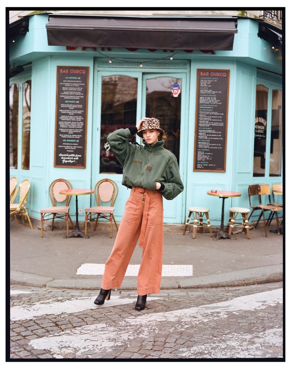 A woman wearing a leopard print hat and a green jacket stands on a street in front of a cafe.