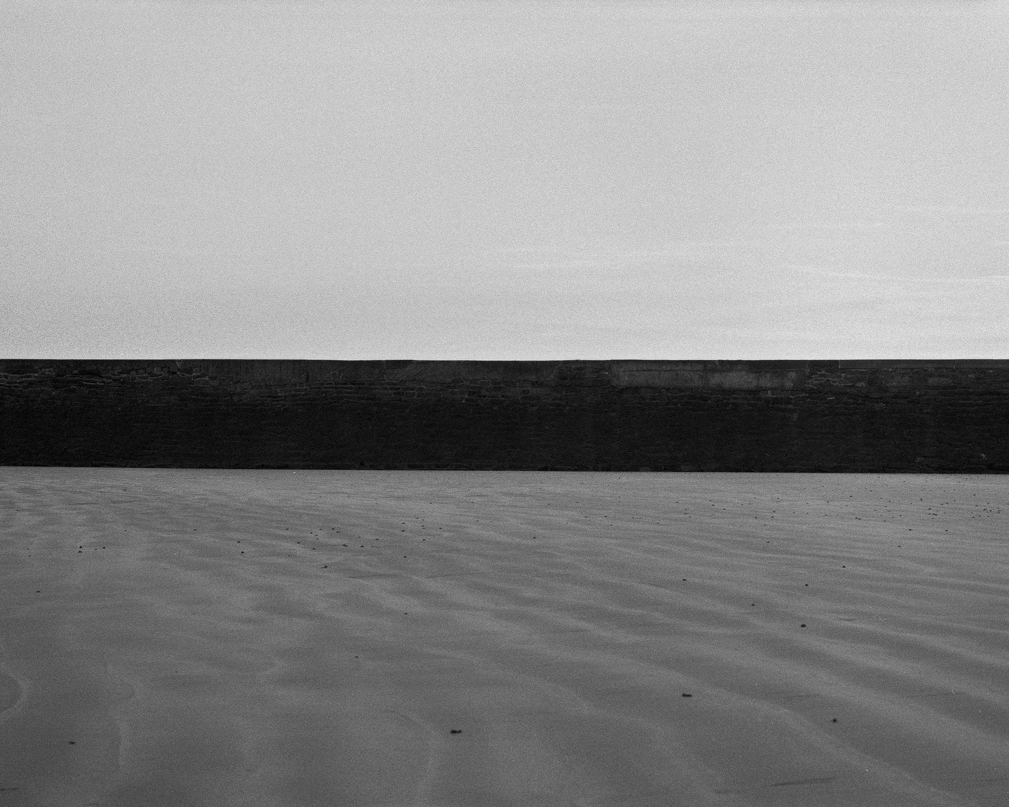 A dark wall separates a light sky from a sandy beach with ripples.
