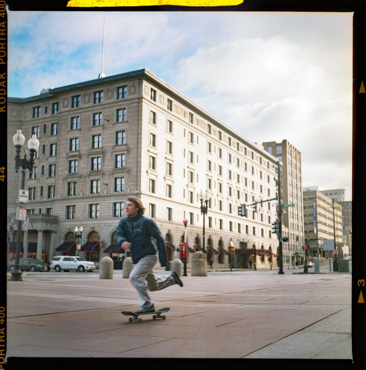 A young man on a skateboard runs across a city square in front of a large building.