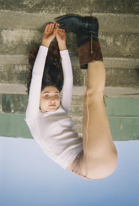 A woman lies on her back with her head near a concrete wall and her legs extended upwards, one foot wearing a cowboy boot.