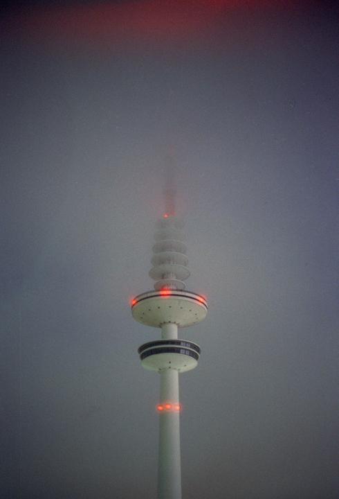 A tall, white television tower with red lights is partially obscured by fog.