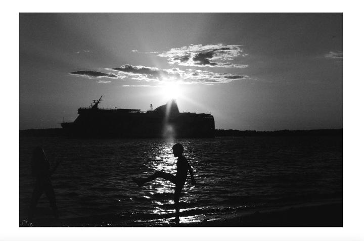 A child stands in the water with a large ship in the background as the sun sets.