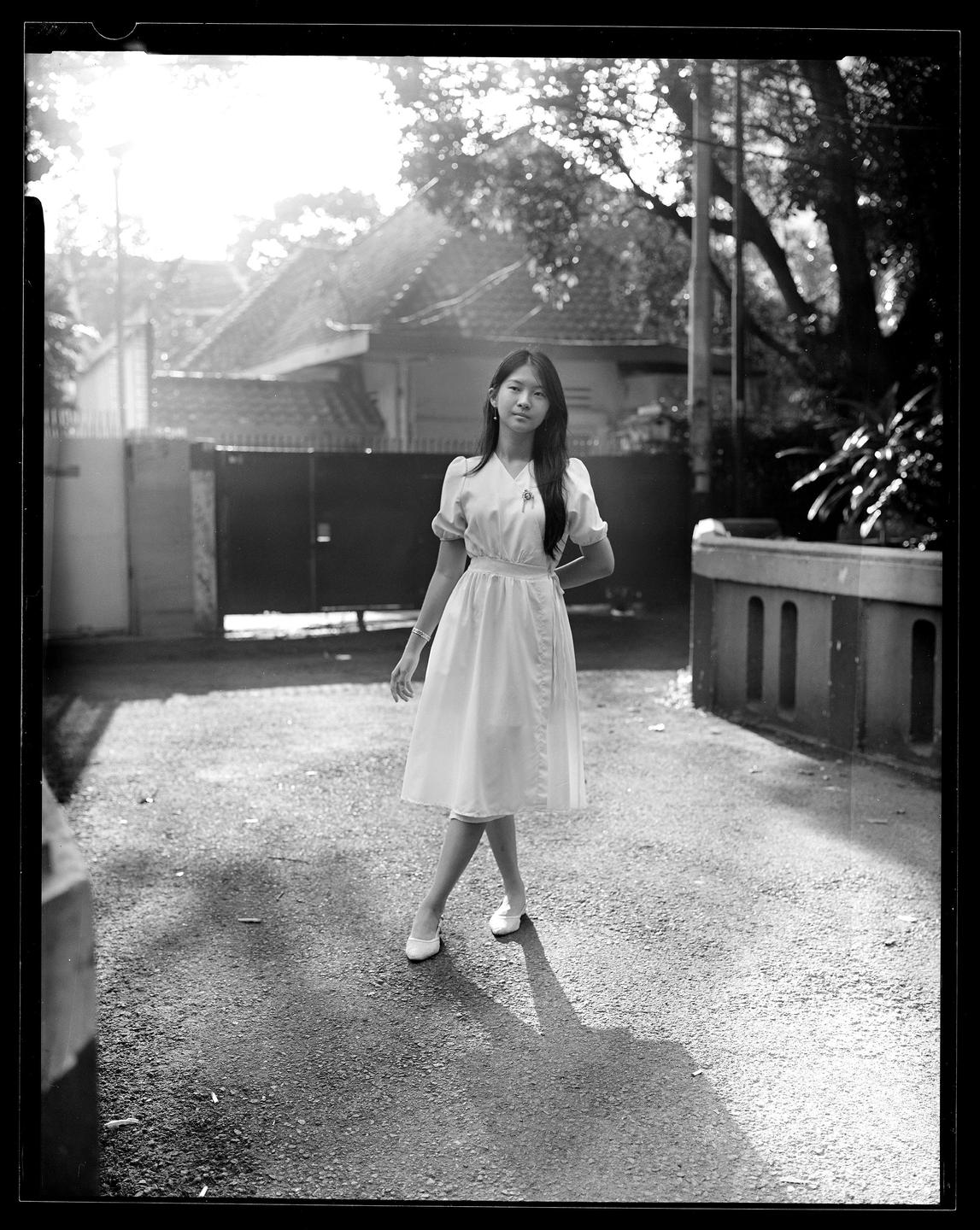 A young woman in a dress stands on a paved surface with houses and trees in the background.