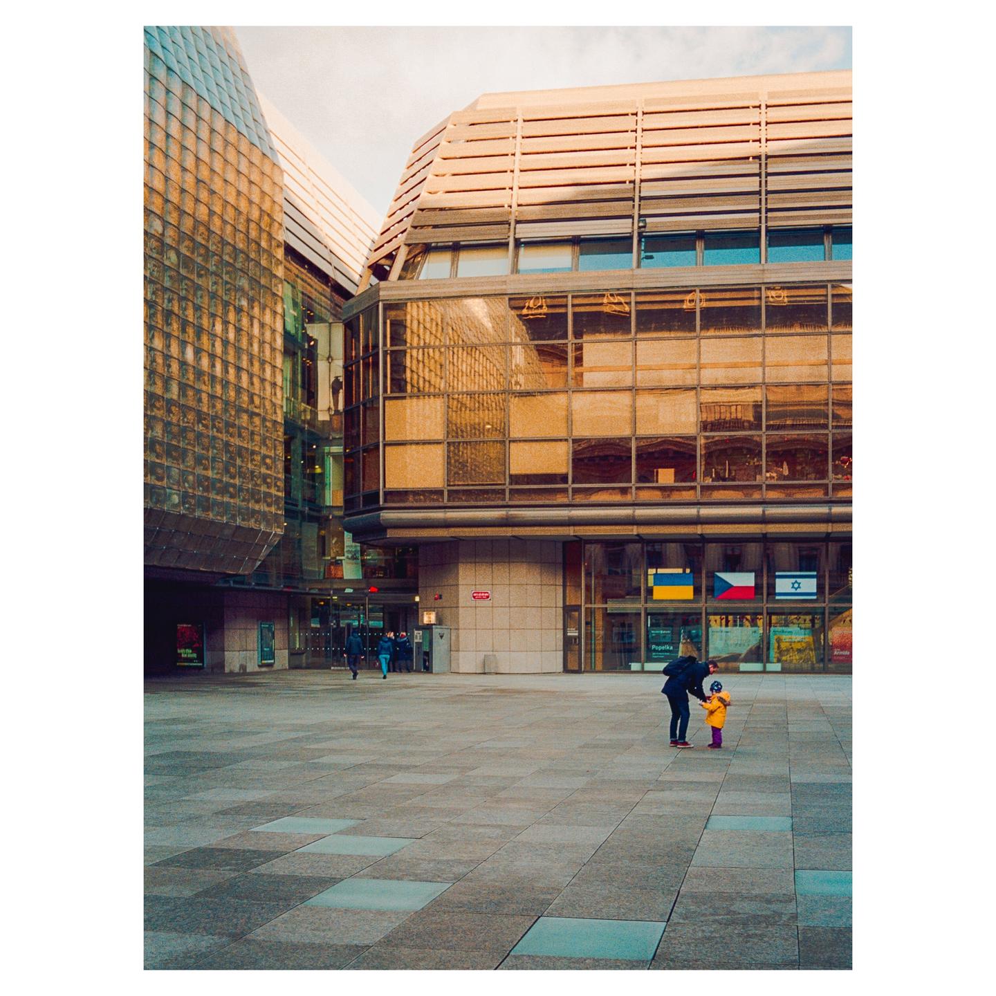 A man bends down to a child in a yellow jacket on a large paved plaza in front of a modern building with reflective windows.