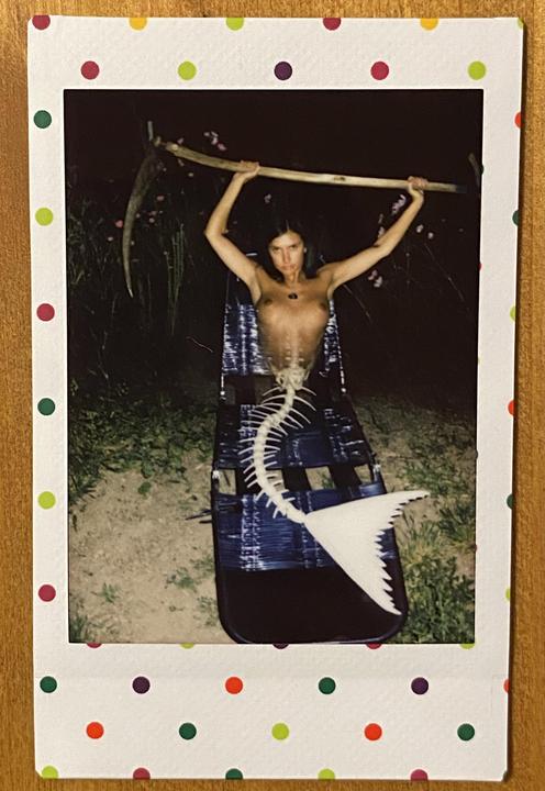A woman with a fish skeleton tail holds a scythe above her head in a night setting.