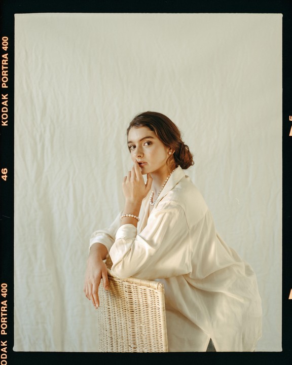 A woman wearing a pearl necklace and bracelet leans on a wicker chair.