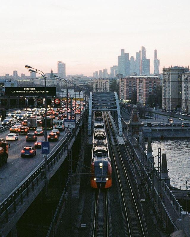 A train travels on tracks next to a busy highway with a city skyline in the background.