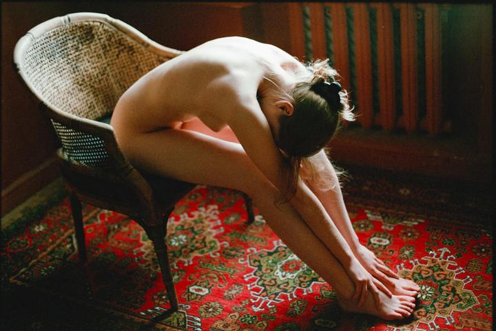 A nude woman sits on a chair with her head bowed to her knees on a patterned rug.