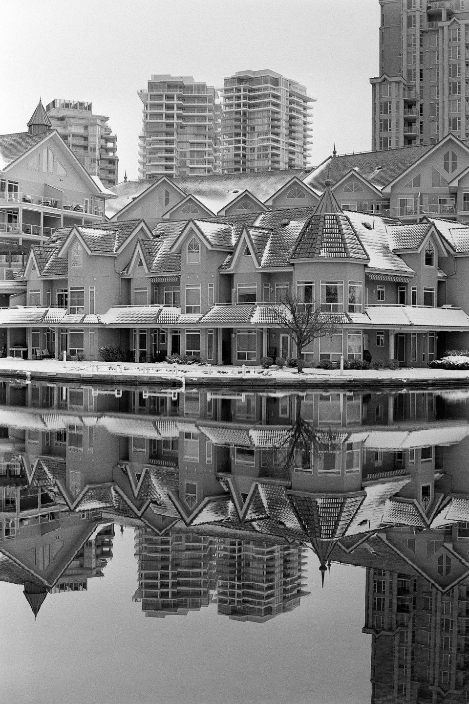 A row of snow-covered houses with pointed roofs is reflected in the water, with tall buildings in the background.