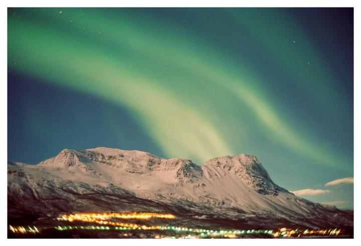 The aurora borealis glows green above a snow-covered mountain and a town at night.