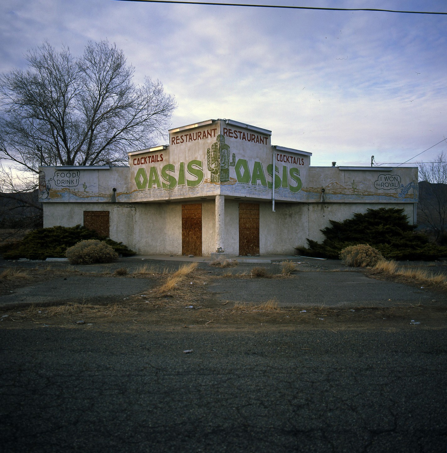An abandoned restaurant with "Oasis" painted on its facade stands under a cloudy sky.