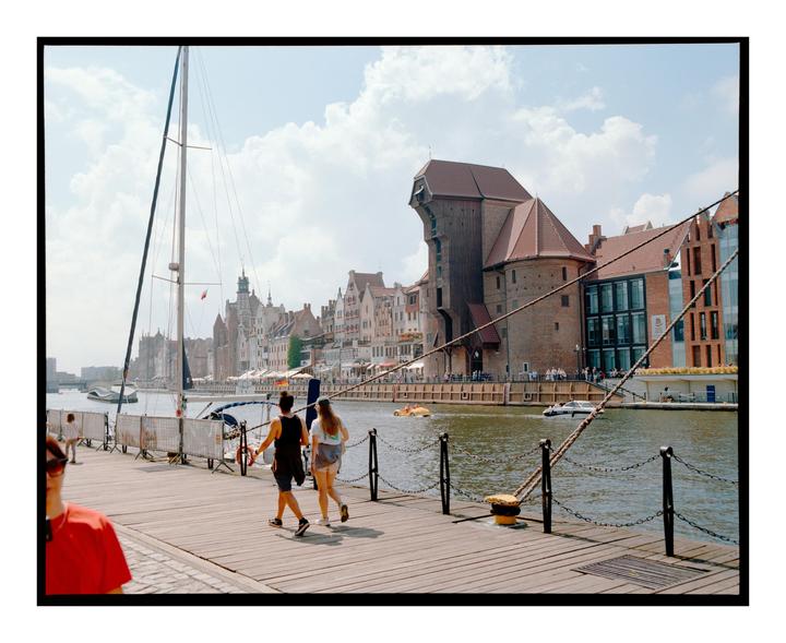 Two women walk along a wooden pier next to a river with historic buildings and a sailboat.
