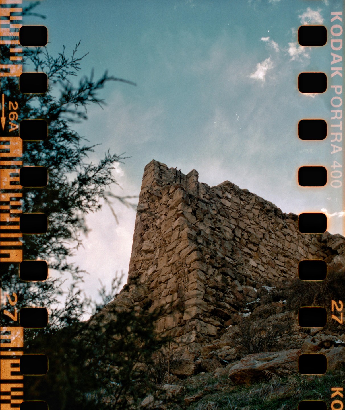 A stone wall of a ruin stands against a blue sky with sparse clouds, framed by dark foliage on the left and bottom.