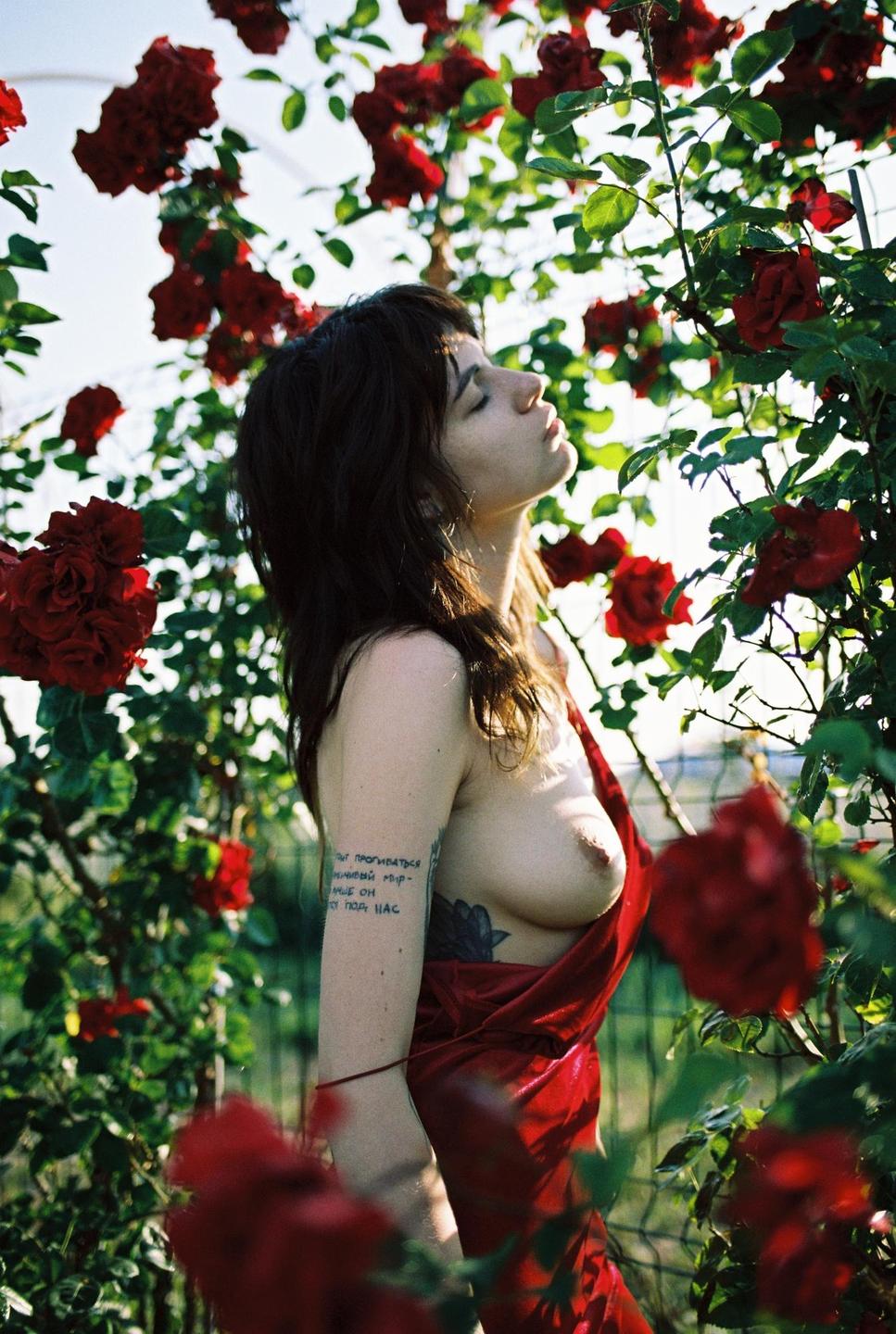 A woman with dark hair and a red dress stands among red roses with her eyes closed and face tilted towards the sky.