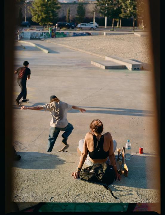 A woman sits on concrete steps watching two men skateboard in a skatepark.