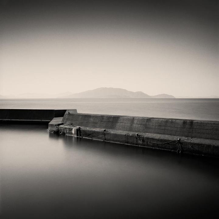 A concrete pier extends into calm water with mountains in the distance under a bright sky.