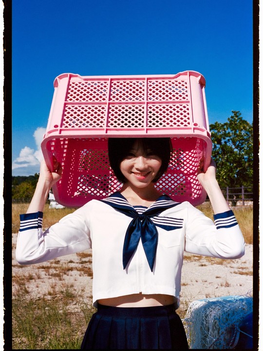 A smiling woman in a sailor uniform holds a pink plastic crate over her head under a clear blue sky.