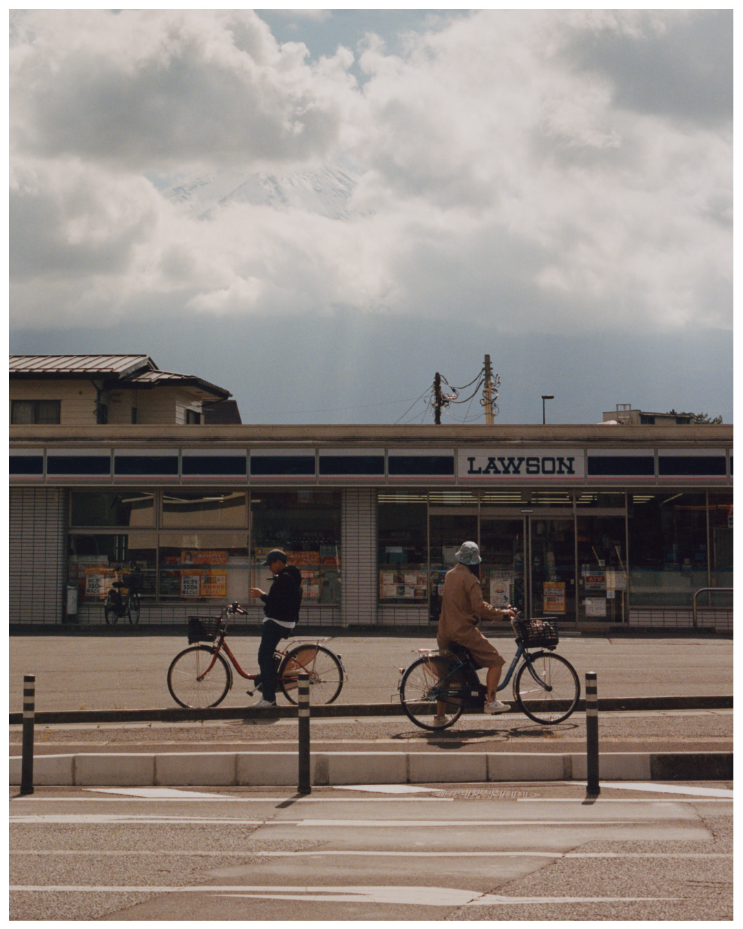 Two people on bicycles are on a street in front of a Lawson convenience store with a snow-capped mountain visible in the distance.