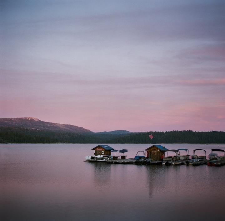 A floating dock with several boats and two small buildings sits on a calm lake surrounded by forested mountains under a pink and purple sky.