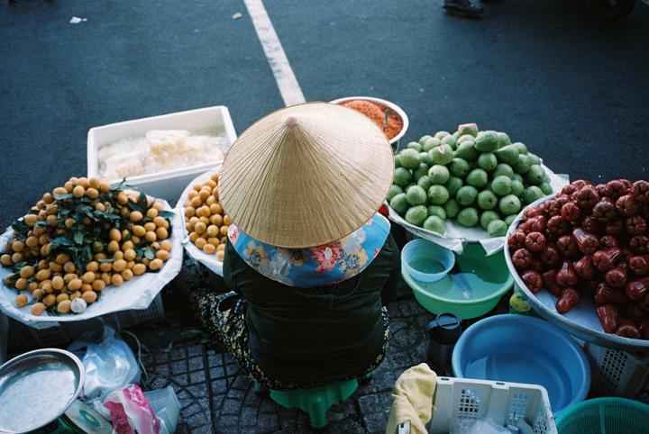 A person wearing a conical hat sits on a stool surrounded by various fruits and containers on a street.