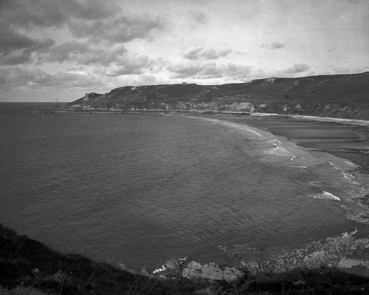 A wide bay with a sandy beach and cliffs is visible under a cloudy sky.