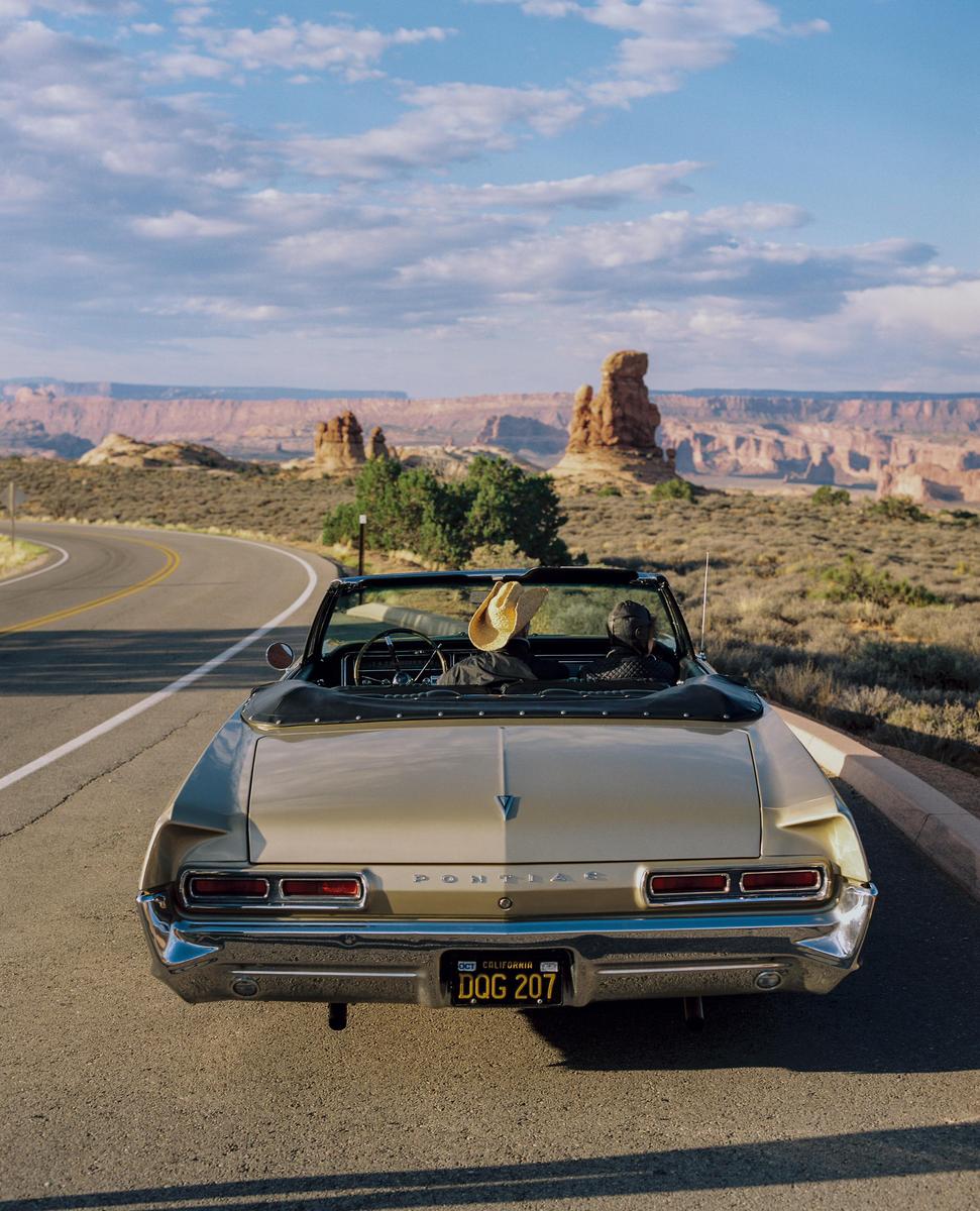 A gold convertible with two people inside is parked on a winding road overlooking a vast desert landscape with rock formations.