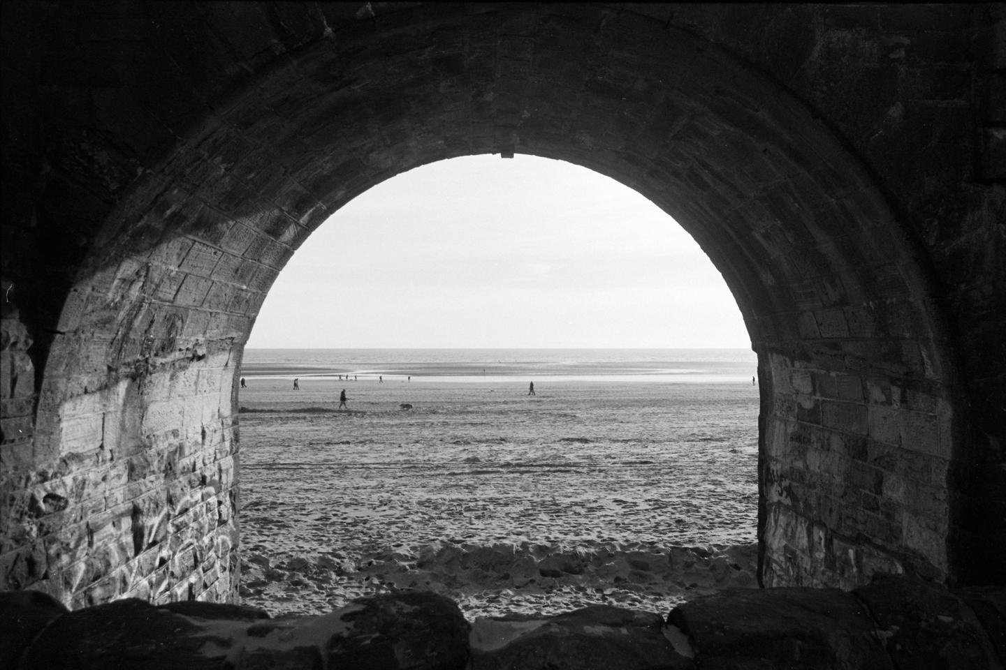 A beach with people and the ocean is visible through a stone archway.