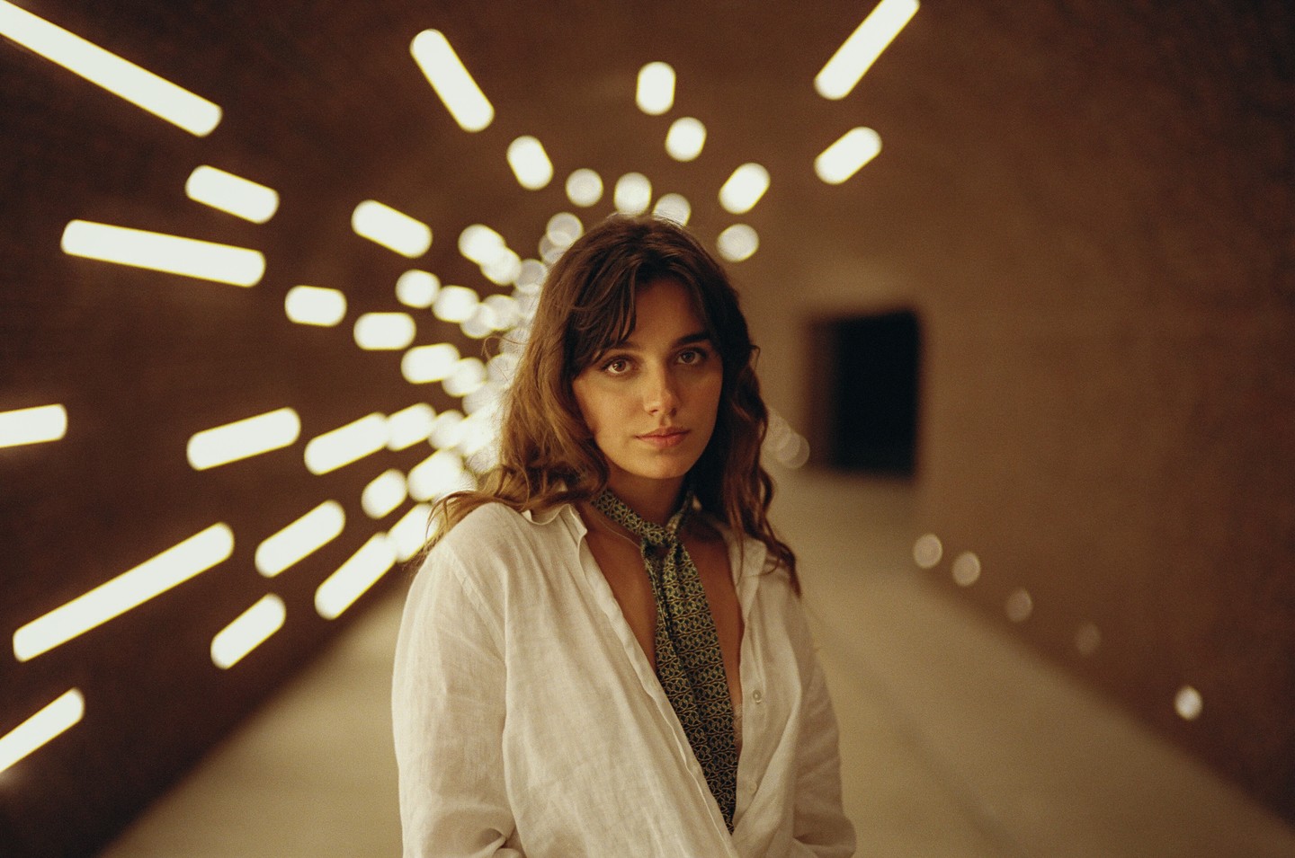 A woman with long hair and a patterned scarf stands in a tunnel with bright, elongated lights on the ceiling.