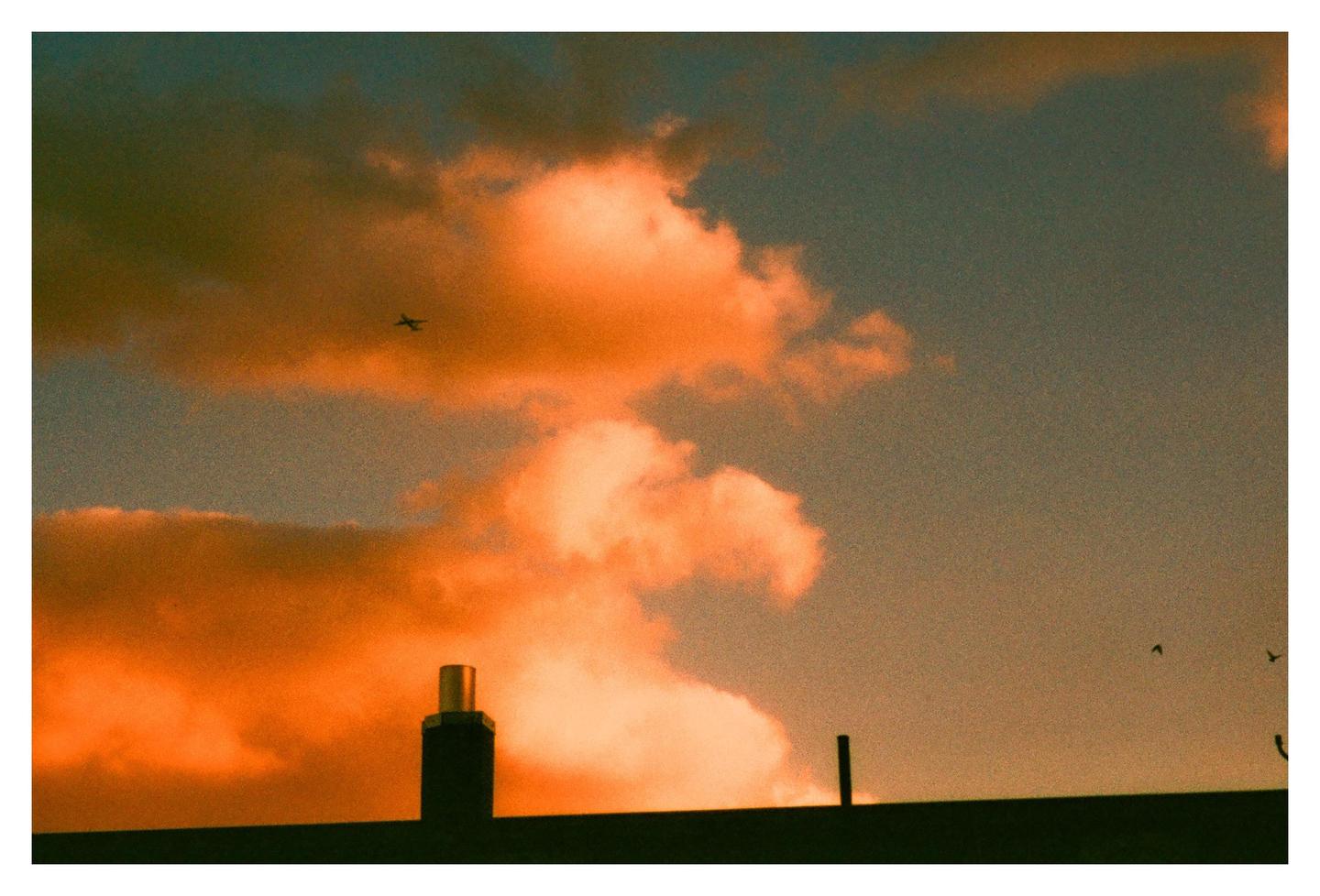 A silhouette of a rooftop with a chimney and an airplane in the distance is visible against a sky filled with orange clouds.