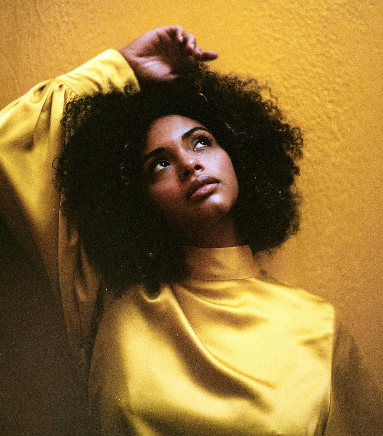 A woman with curly hair and a yellow top looks up with her arm raised against a yellow background.