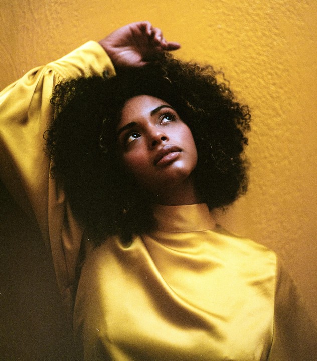 A woman with curly hair and a yellow top looks up with her arm raised against a yellow background.