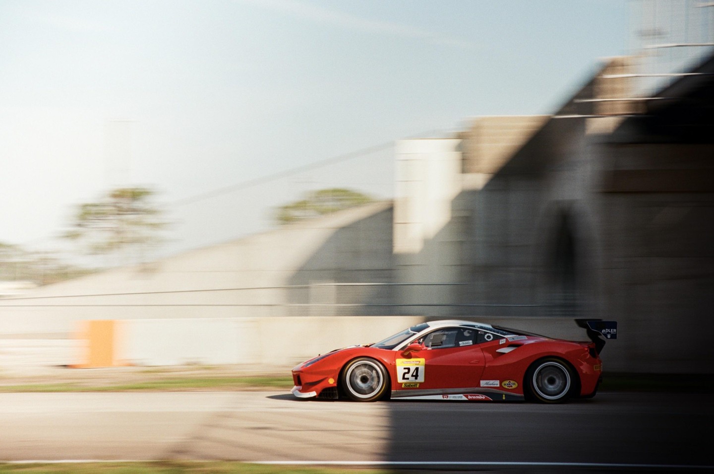 A red race car with the number 24 on its side speeds past a concrete structure, with motion blur in the background.