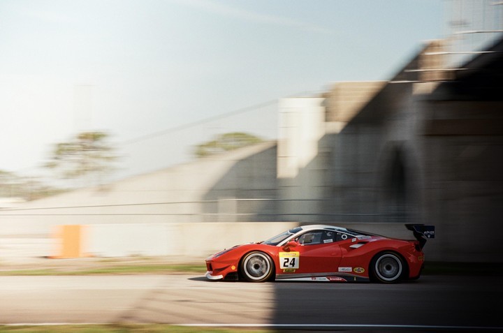 A red race car with the number 24 on its side speeds past a concrete structure, with motion blur in the background.