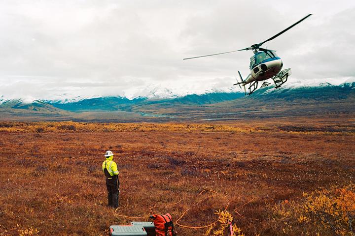 A helicopter approaches a person standing in a vast, autumn-colored landscape with snow-capped mountains in the distance.