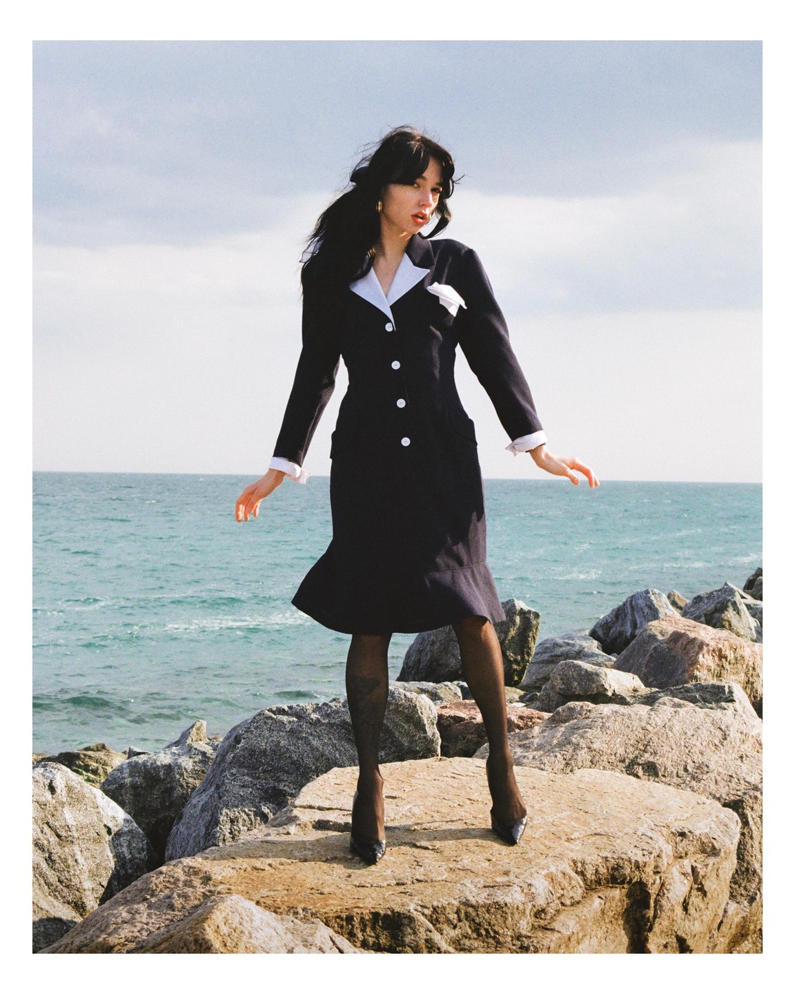 A woman in a dark dress with white accents stands on rocks by the sea.