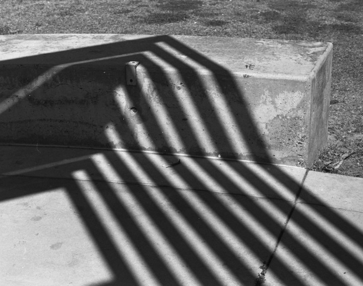 Strong shadows from a fence fall across a concrete bench and the ground next to a patch of grass.