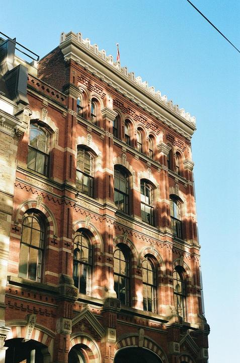 A red brick building with many arched windows is illuminated by sunlight and shadows.