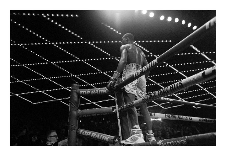 A boxer stands in a boxing ring, looking out at the crowd and the illuminated ceiling.
