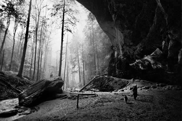 A person and a dog walk along a path next to a large rock formation and a forest.
