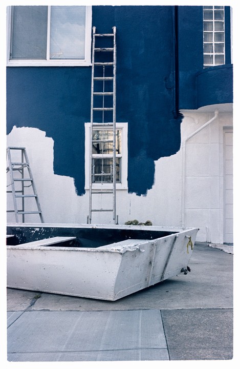 A boat sits on the ground next to a building being painted blue and white, with ladders leaning against the wall.