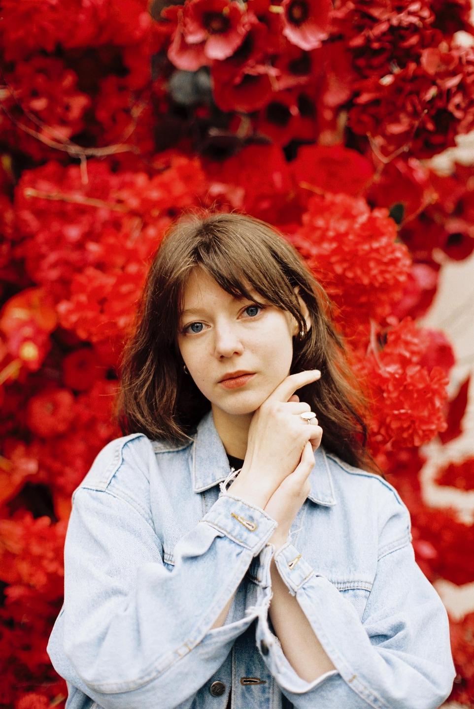 A woman with brown hair and a denim jacket poses in front of a wall of red flowers.