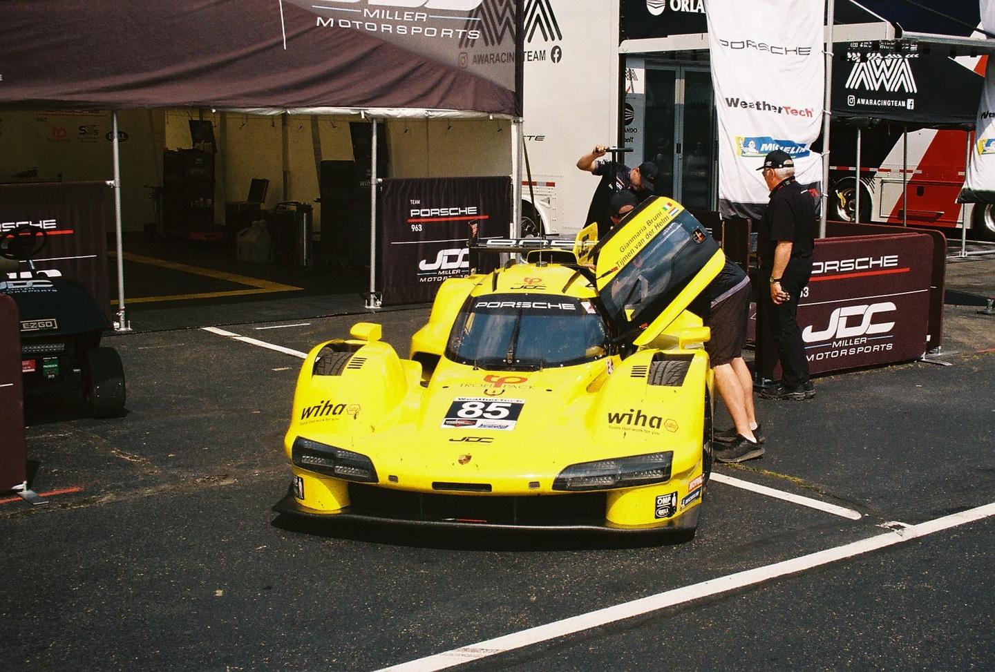 A yellow race car is parked on asphalt with several men standing around it and tents in the background.