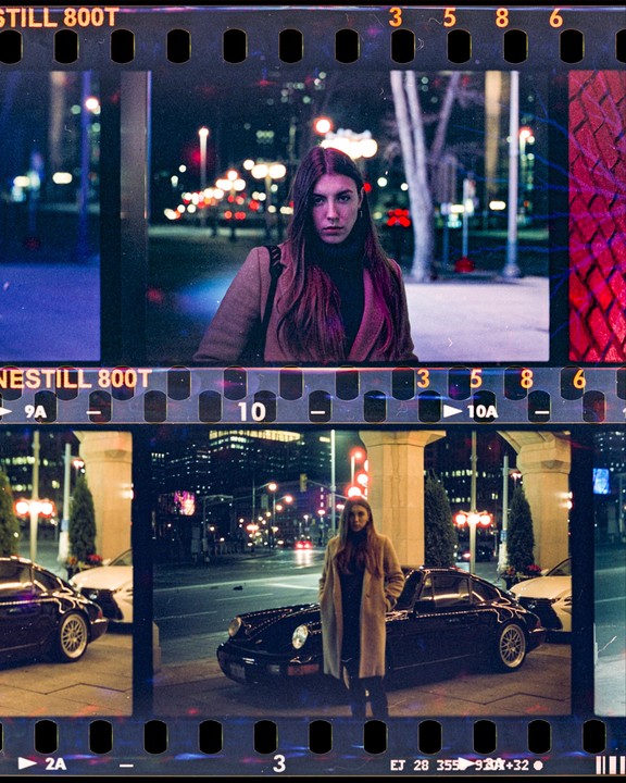 A woman stands on a city street at night, with a car parked nearby.