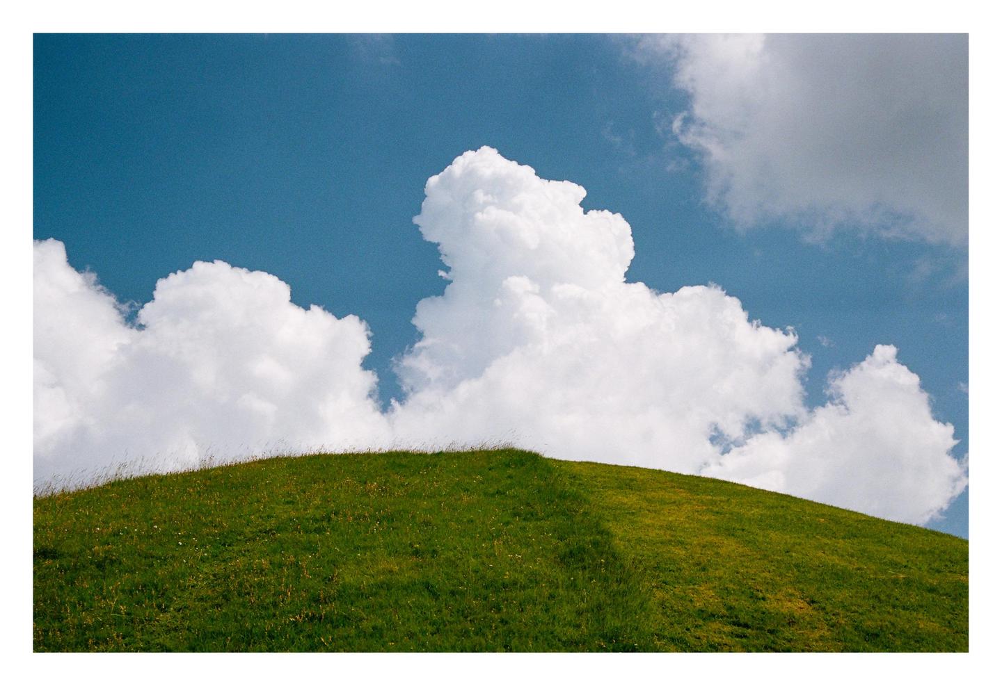 A green grassy hill is topped with large white cumulus clouds against a blue sky.