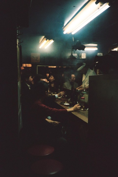 People sit at a bar counter in a dimly lit establishment with fluorescent lights on the ceiling.